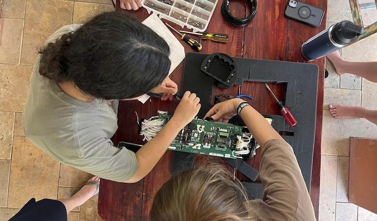 两个学生在电脑板上工作. 桌子周围放着螺丝刀、电缆和一个工具箱.