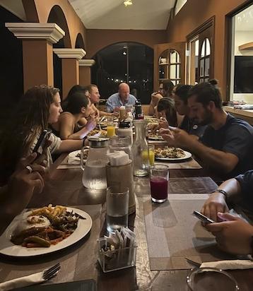 学生和研究人员坐在一张长桌旁，面前摆着装满食物的餐盘. 一些人在吃东西，另一些人在互相交谈.